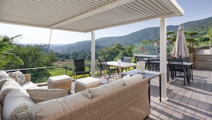 A covered outdoor seating area with wicker furniture, a table, and chairs, overlooking a scenic landscape.
