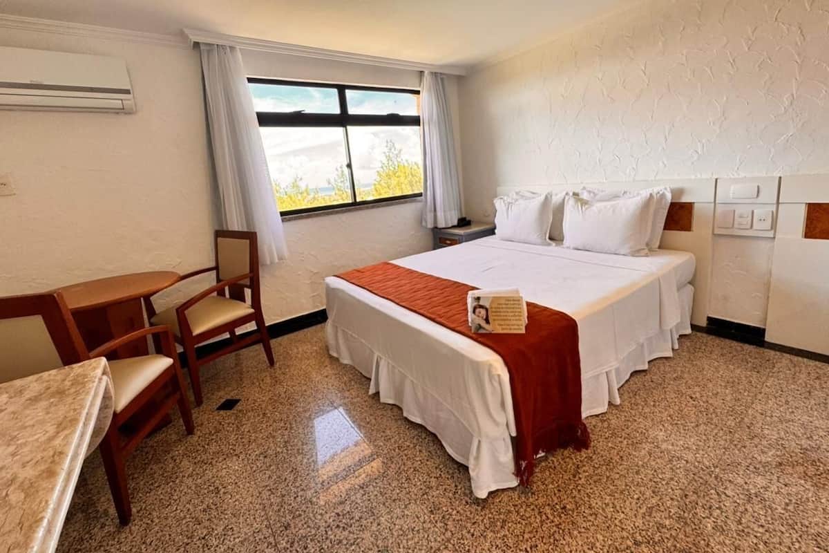 A hotel room with a bed, a desk, chairs, a window with curtains, and an air conditioning unit.