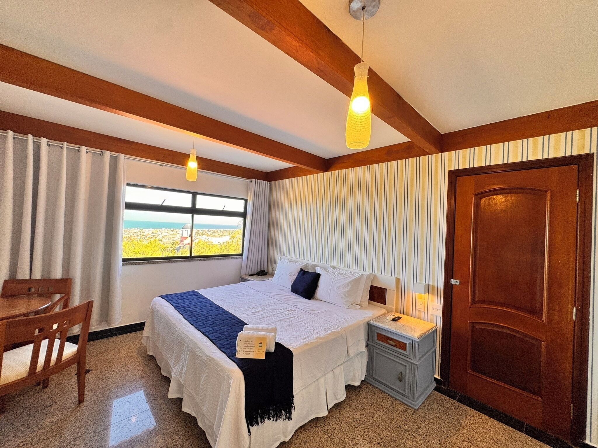 A hotel room with a bed, bedside table, nightstand, wooden chair, and a window with a view.