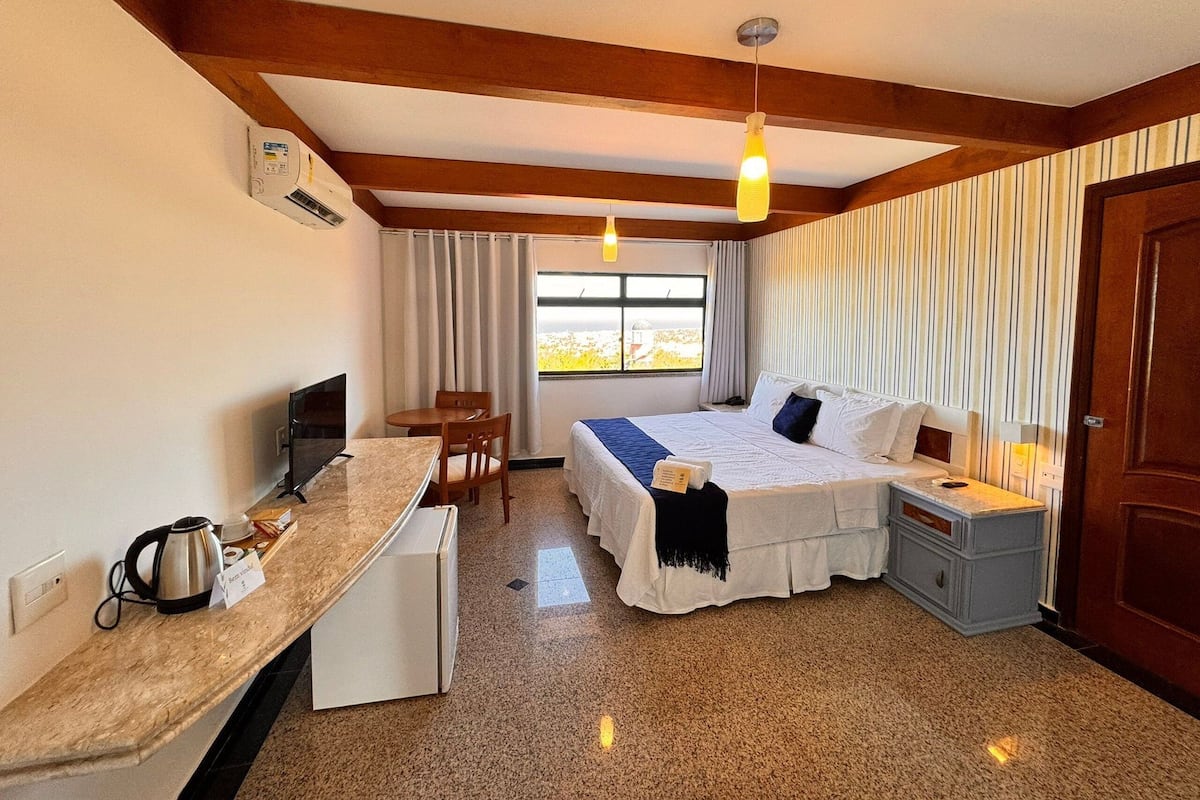 A hotel room with a bed, a desk, a chair, a small refrigerator, a window with curtains, and a door.