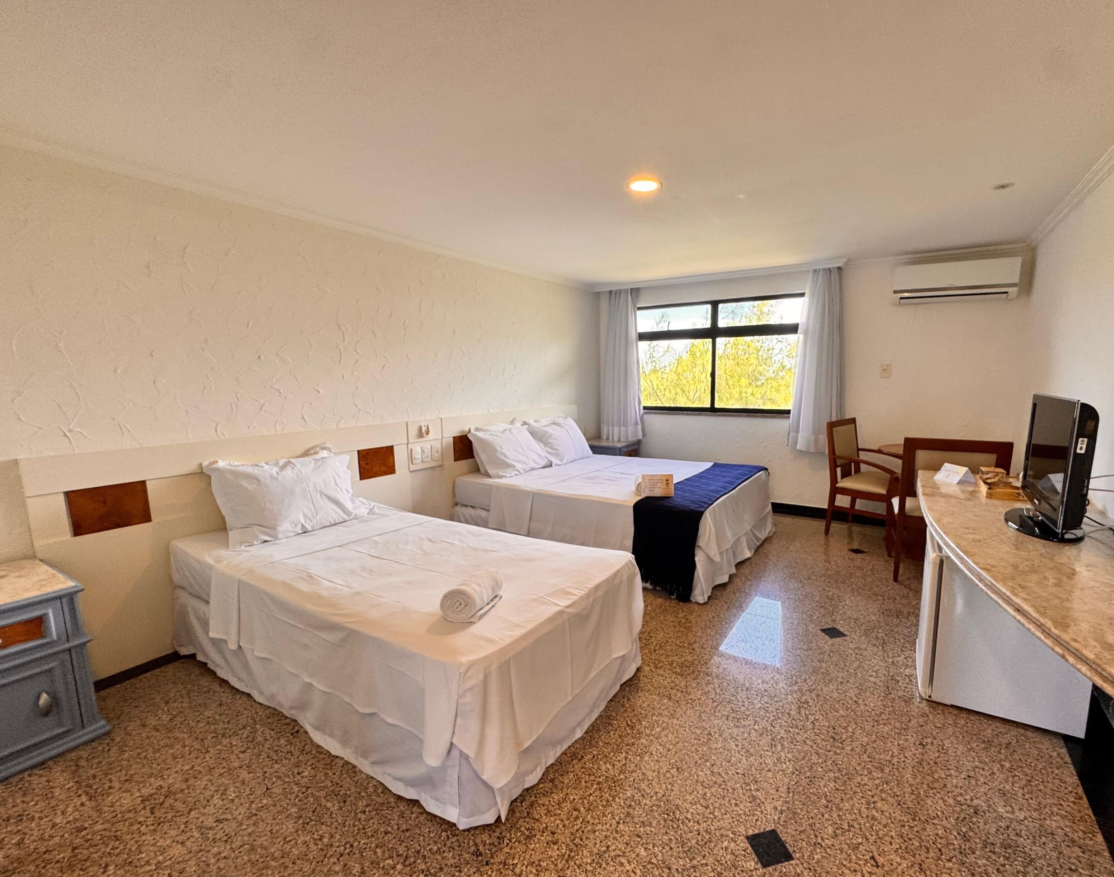 A hotel room with two beds, a desk, a chair, a television, and a window with a view of trees.
