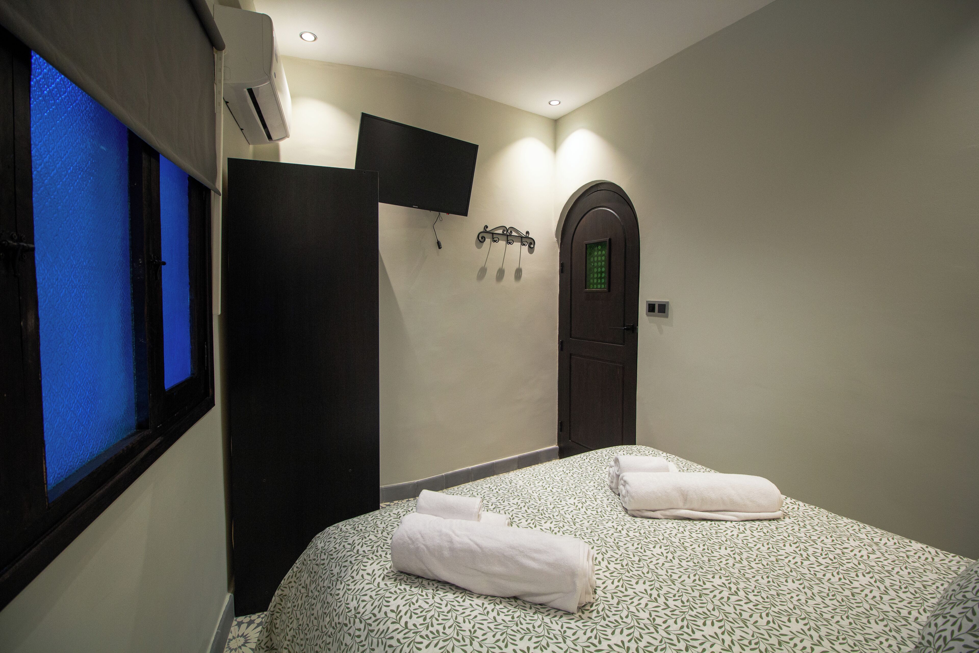 A hotel room with a bed, two towels, a TV, an air conditioning unit, and a door with a green light.