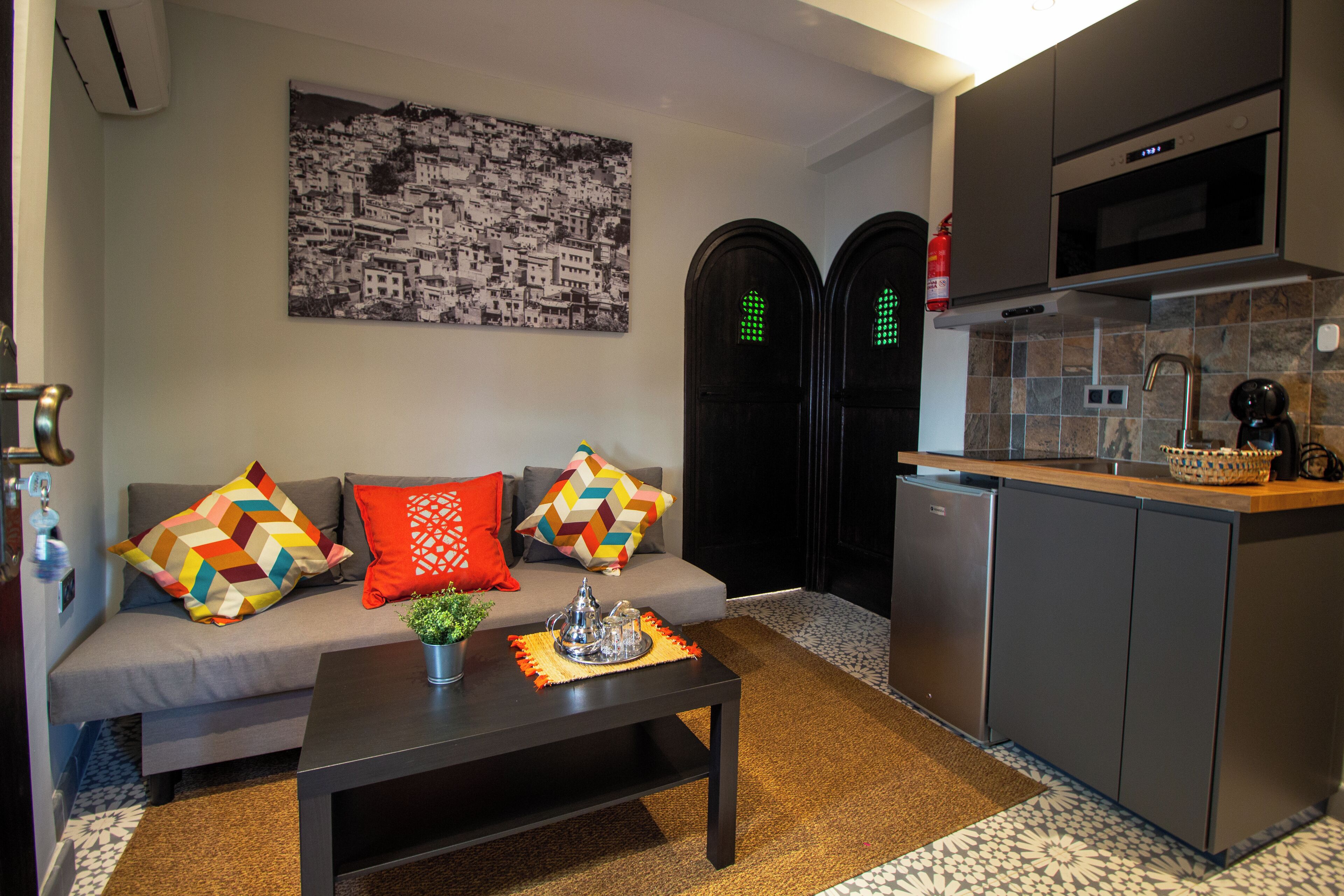 Un moderno espacio de sala con un sofá, cojines coloridos, una mesa de centro con un objeto decorativo, una cocina con microondas y máquina de café, y un cuadro de un paisaje urbano en blanco y negro en la pared.