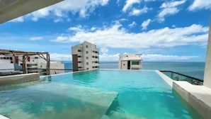 Uma piscina infinita com águas cristalinas, vista para o oceano e edifícios ao fundo.