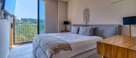 A bedroom with a large bed, a bedside table, a lamp, a wall-mounted dreamcatcher, and a balcony with a view of buildings and greenery.