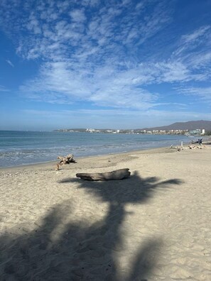Una playa con un tronco grande, cielo despejado y edificios a lo lejos.