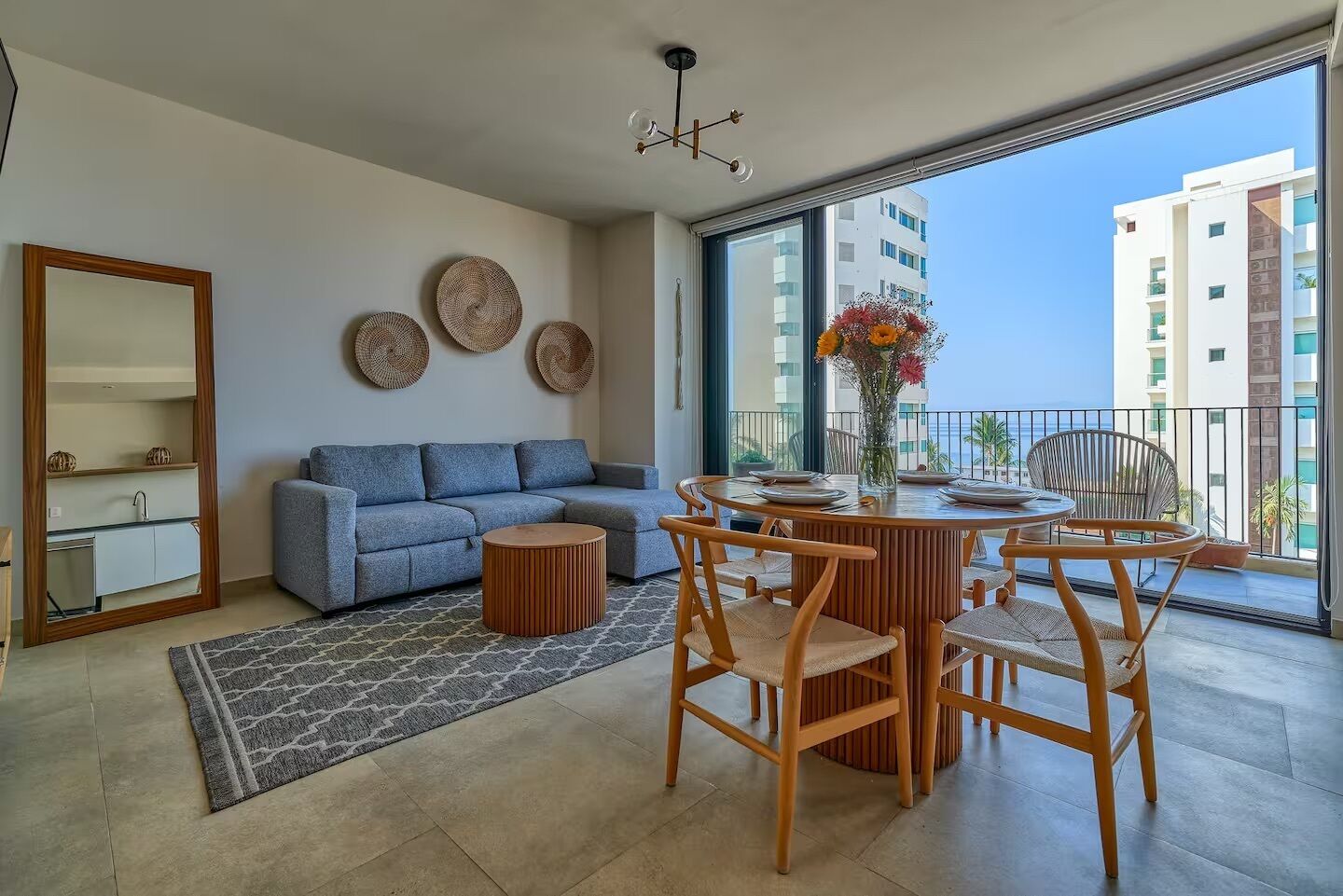 A modern living room with a sofa, round wooden table, and chairs, a small rug, and a large window with a view of buildings and the sea.