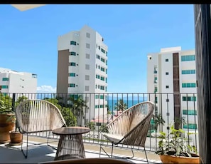 Uma varanda com cadeiras de vime e uma pequena mesa, com vista para uma paisagem urbana com edifícios altos e céu claro.