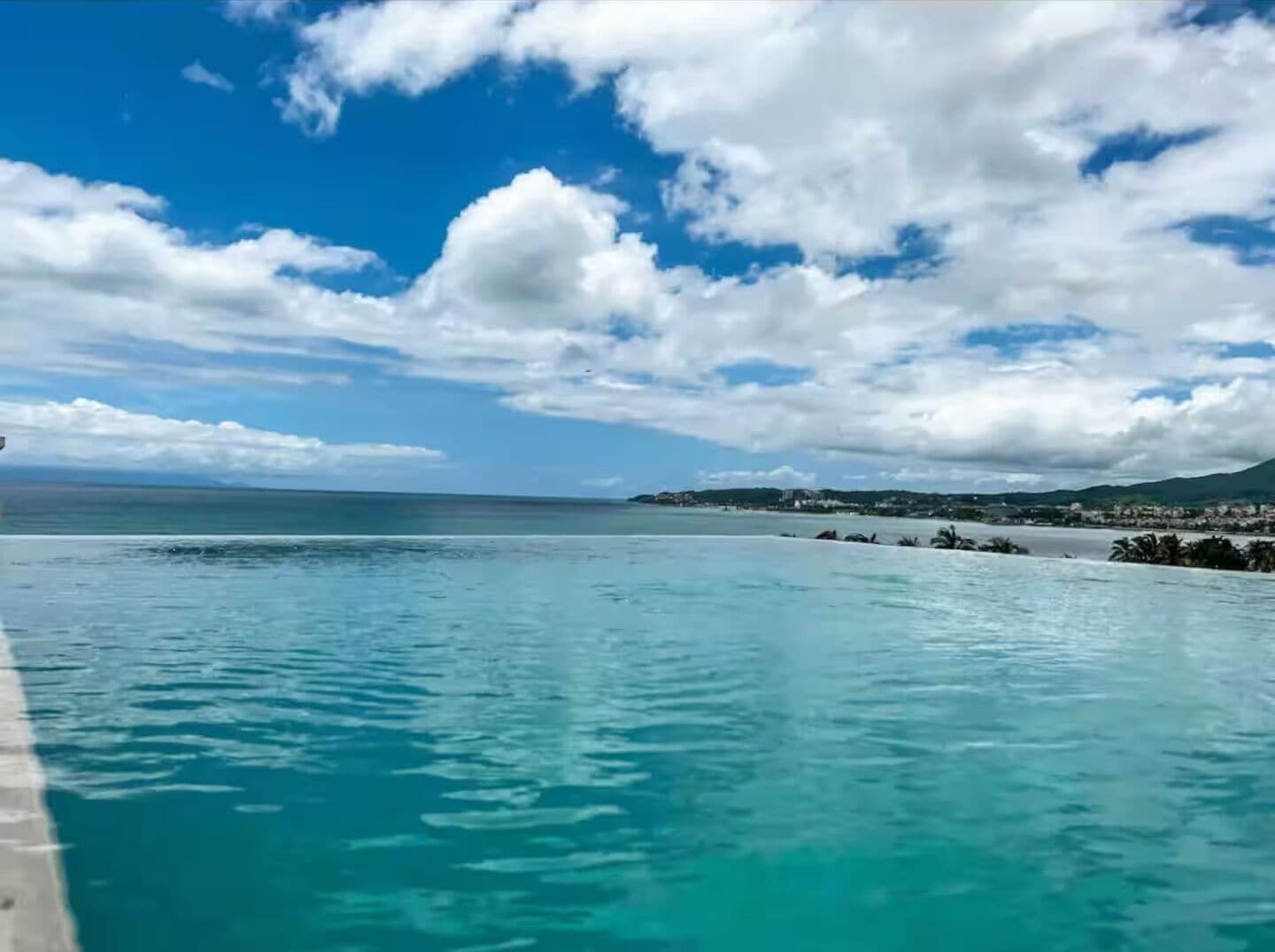 Klar udsigt over en swimmingpool med en fjerntliggende kystlinje og et delvist overskyet himmelbillede.