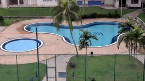 A swimming pool with a palm tree in the foreground, surrounded by a fenced area and a building with balconies in the background. - Homestay Ziyan with pool near beach (Port Dickson Town)