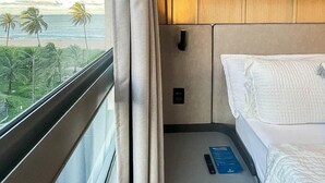 Een hotelkamer met een bed, nachtkastje, raam met gordijnen en uitzicht op de oceaan en palmbomen.