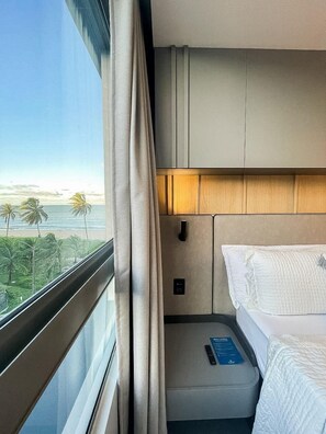 Ein Hotelzimmer mit Bett, Nachttisch, einem Fenster mit Vorhängen und Blick auf das Meer und Palmen.