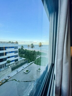 Blick aus einem Fenster auf ein Gebäude mit Balkon, gepflastertem Bereich und Palmen.