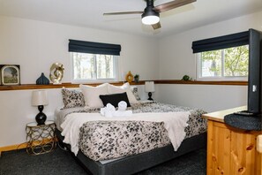 Une chambre à coucher comprenant un lit, deux tables de chevet, un ventilateur de plafond et une fenêtre donnant sur des arbres.
