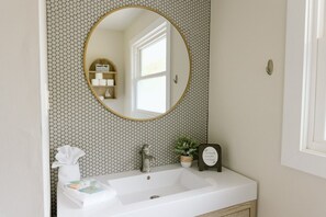 Une salle de bain avec un lavabo blanc, un miroir rond doté d’un cadre doré, une petite plante en pot, un distributeur de savon et une serviette.
