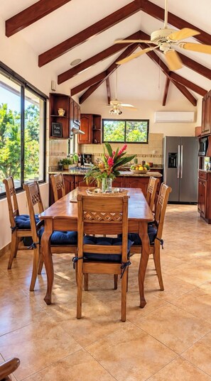 Een ruime keuken met een hoge plafond en zichtbare balken, een eettafel met stoelen en een groot raam dat uitzicht biedt op groen buiten.