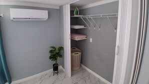 Une salle de bain avec un climatiseur blanc, une plante en pot, un étendoir suspendu et une poubelle.