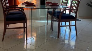 Uma sala de jantar com uma mesa de vidro, quatro cadeiras de madeira, um vaso com planta e uma janela com cortinas.