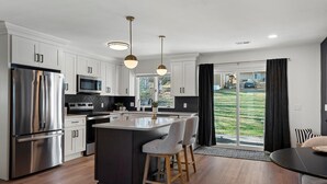 A modern kitchen with stainless steel appliances, a central island with bar stools, pendant lighting, and a view of a backyard through large sliding doors. - Top-Rated Premier Modern Getaway (Petersburg)