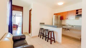 A modern kitchen with a refrigerator, sink, and bar stools. There is a leather couch in the living area.