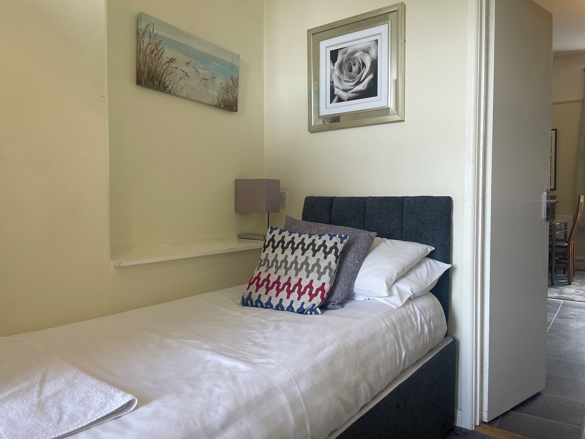 A single bed with a grey headboard, white bedding, and a patterned pillow. There is a wall-mounted lamp and a framed picture above the bed.