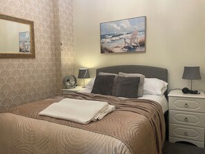 Ein ordentlich bezogenes Bett mit gefalteter Decke, ein Nachttisch mit Lampe, ein weiterer Nachttisch mit Ventilator, eine Kommode mit Schubladen und ein Bild einer Strandlandschaft an der Wand.