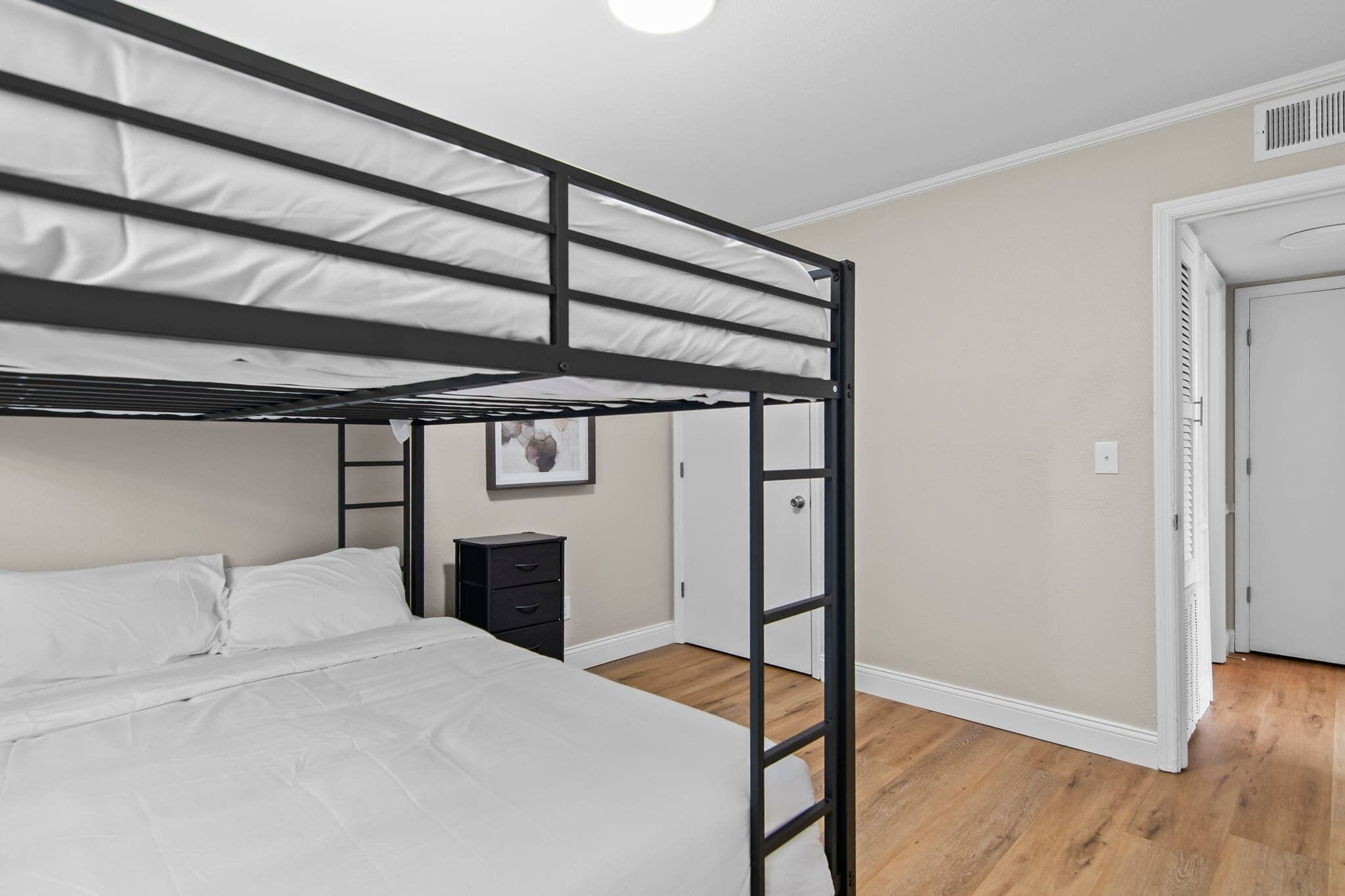 Habitación con literas, suelo de madera, una mesita de noche pequeña y una puerta.