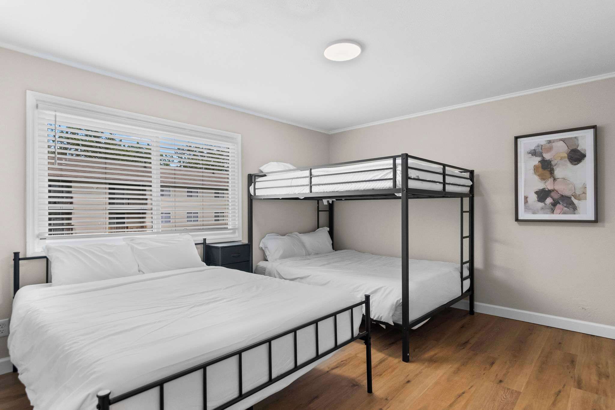 A room with a bunk bed, a bedside table, a window with blinds, and a framed picture on the wall.
