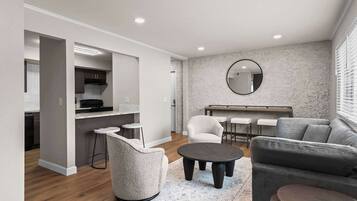 A modern living room with a grey sofa, a round coffee table, and a white armchair. There is a kitchen area with dark cabinets and a black range hood. A round mirror hangs on the wall, and a dining table with four chairs is visible.