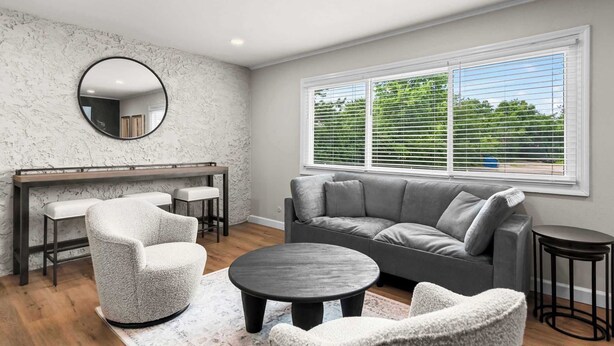 A living room with a gray sofa, round coffee table, and two armchairs. There is a round mirror on the wall and a window with blinds. - Newly Renovated Unit in Central Spfld! #203 (Springfield)