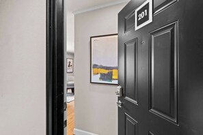 Un couloir avec une porte noire portant le numéro « 201 », un tableau abstrait encadré, et un aperçu d’une pièce où l’on aperçoit une chaise blanche.