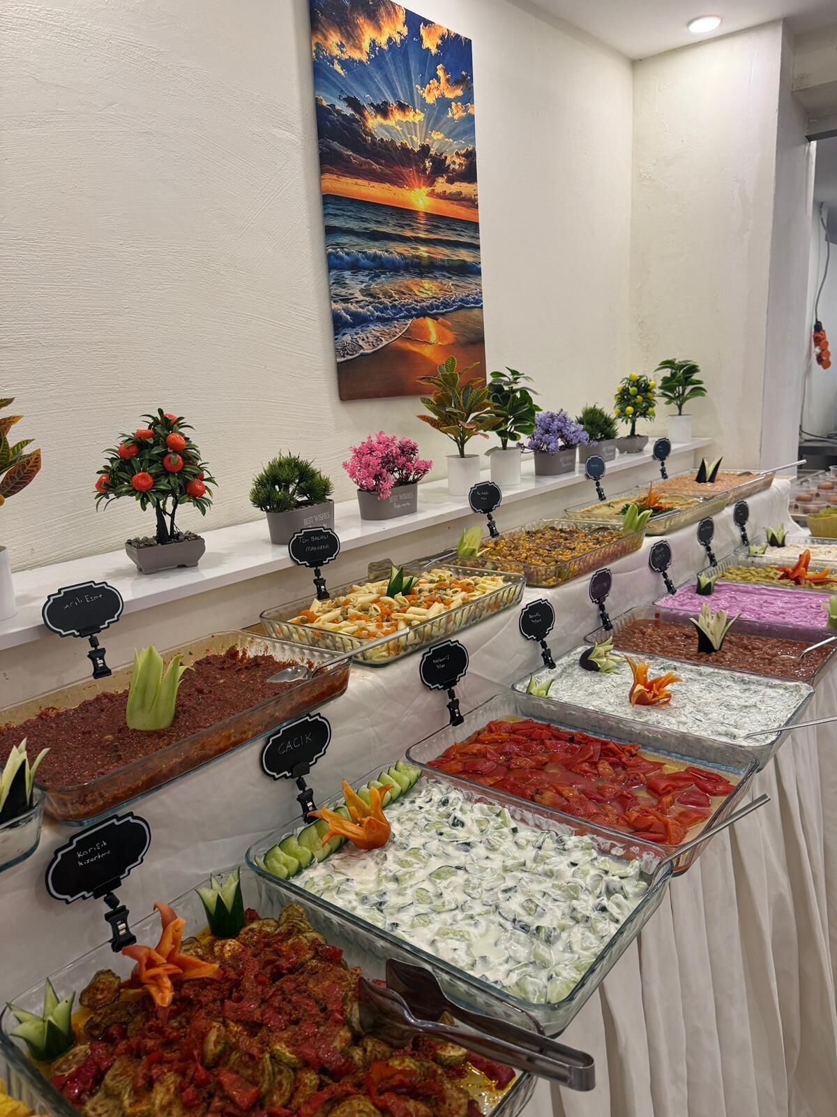 Ein Buffet mit verschiedenen Speisen, ein Sonnenuntergangsgemälde und Zimmerpflanzen.