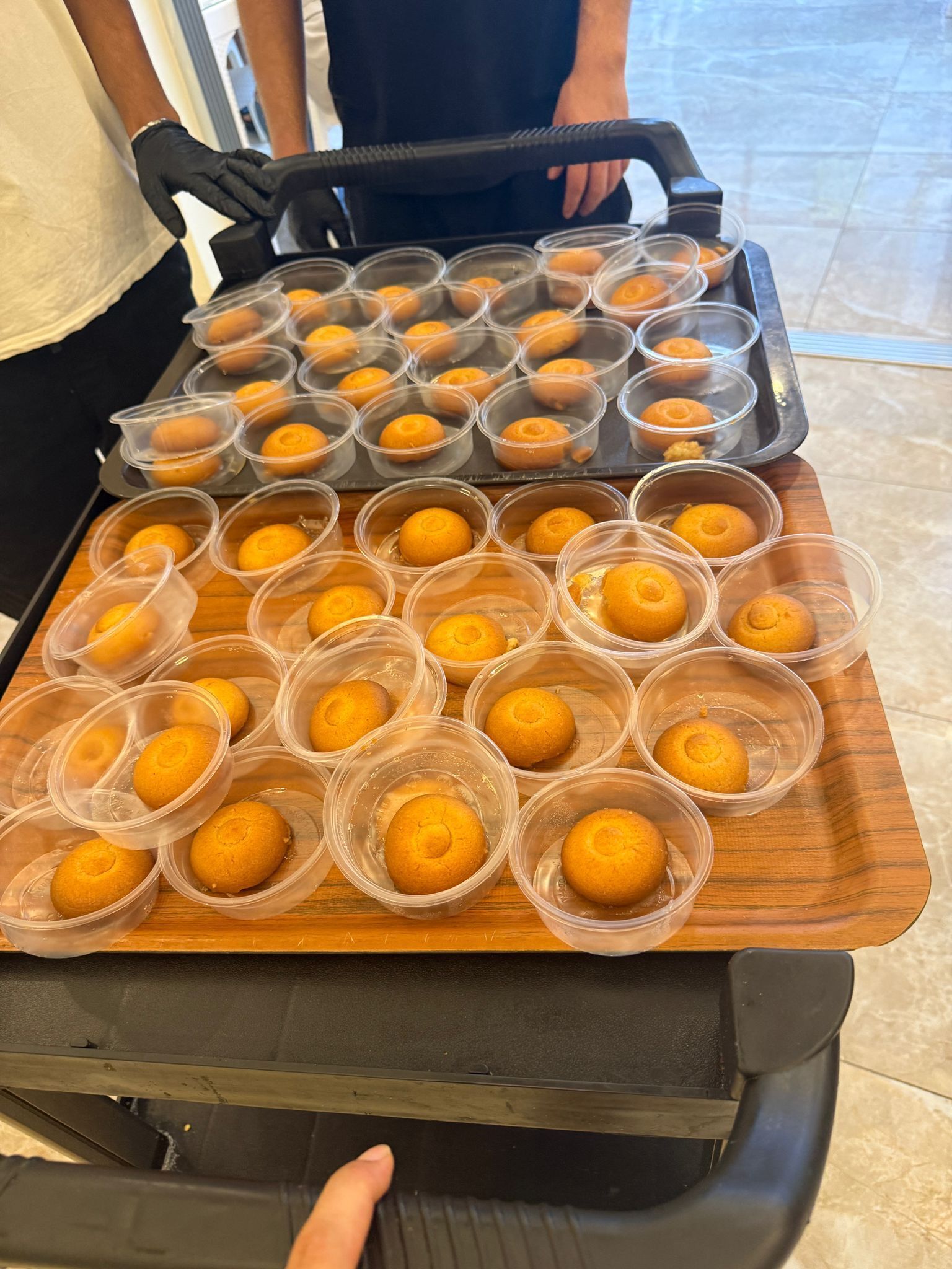 A tray with multiple plastic containers holding round, golden-brown baked goods, likely muffins or cakes, on a wooden surface.