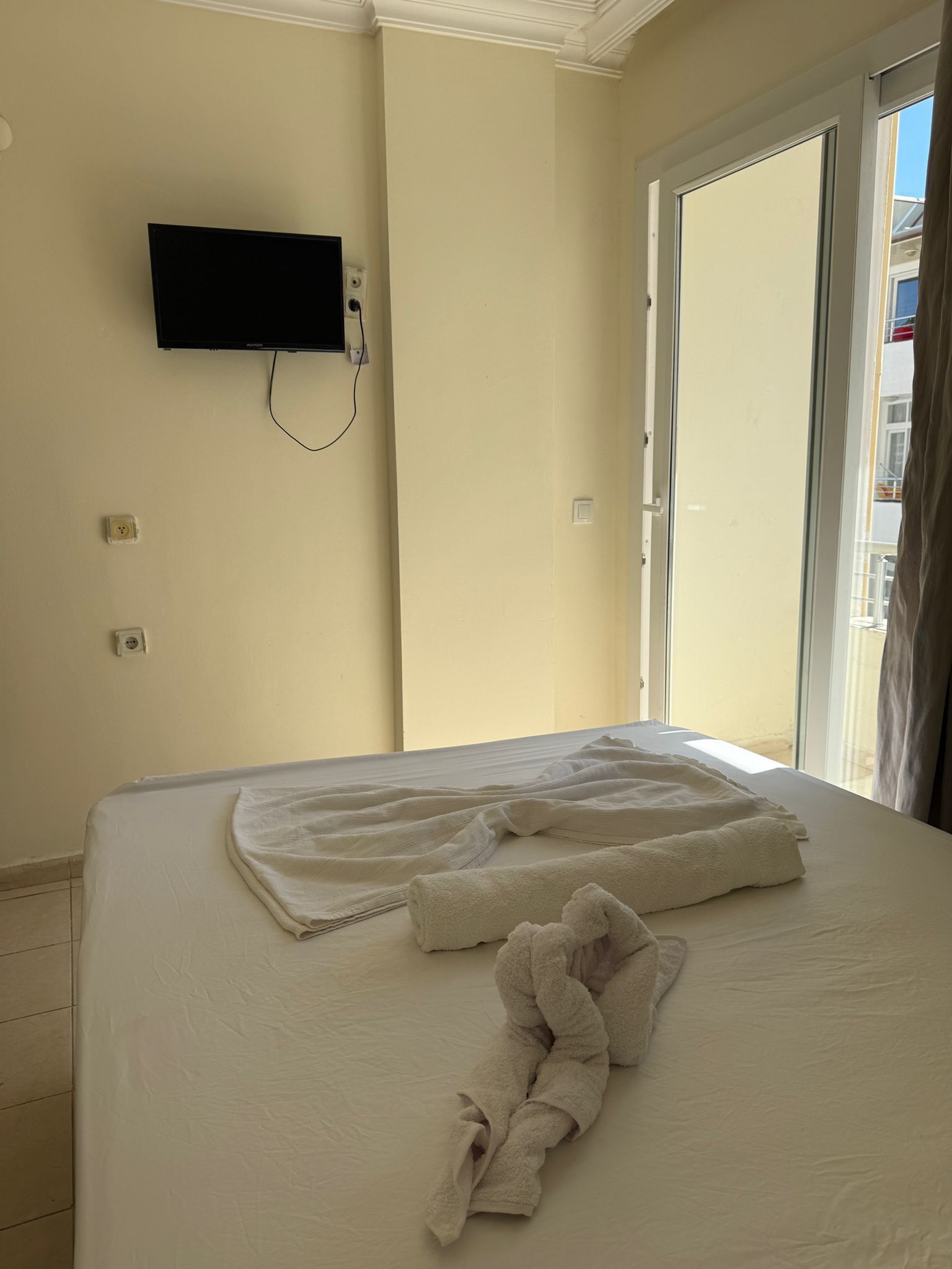 A hotel room with a bed, a flat-screen TV mounted on the wall, and a door leading to an outdoor balcony.