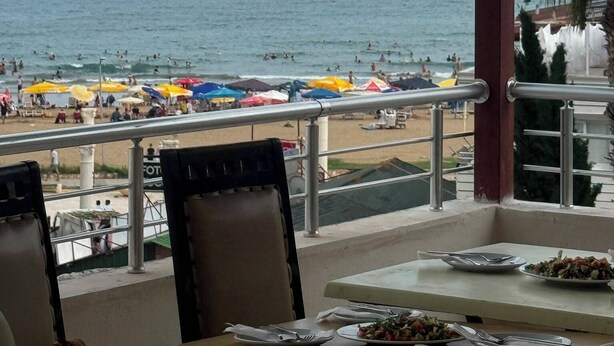 A seaside view from a covered outdoor area with a table set for dining, overlooking a beach with umbrellas and people. - Holiday Hotel (Erdemli)