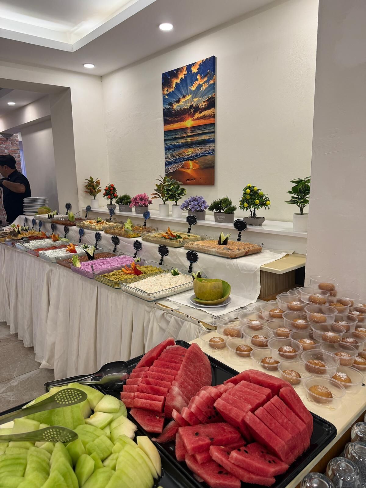 A buffet with various food items, including watermelon and other fruits, and a painting of a sunset on the wall.