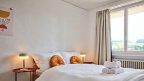 A neatly made bed with white linens, two bedside lamps, and a view of a green landscape through a window with curtains. - Nature & tranquillity I Bern I Family I Parking free I Pet-friendly (Köniz)