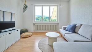 A modern living room with a flat-screen TV, a sofa, a round coffee table, and a large window with a view of greenery. - Nature & tranquillity I Bern I Family I Parking free I Pet-friendly (Köniz)