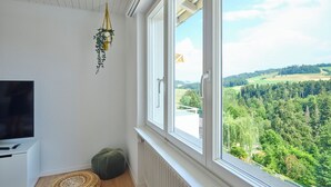 A room with a TV, a woven rug, a green pouf, and a view of a forest through a window. - Nature & tranquillity I Bern I Family I Parking free I Pet-friendly (Köniz)