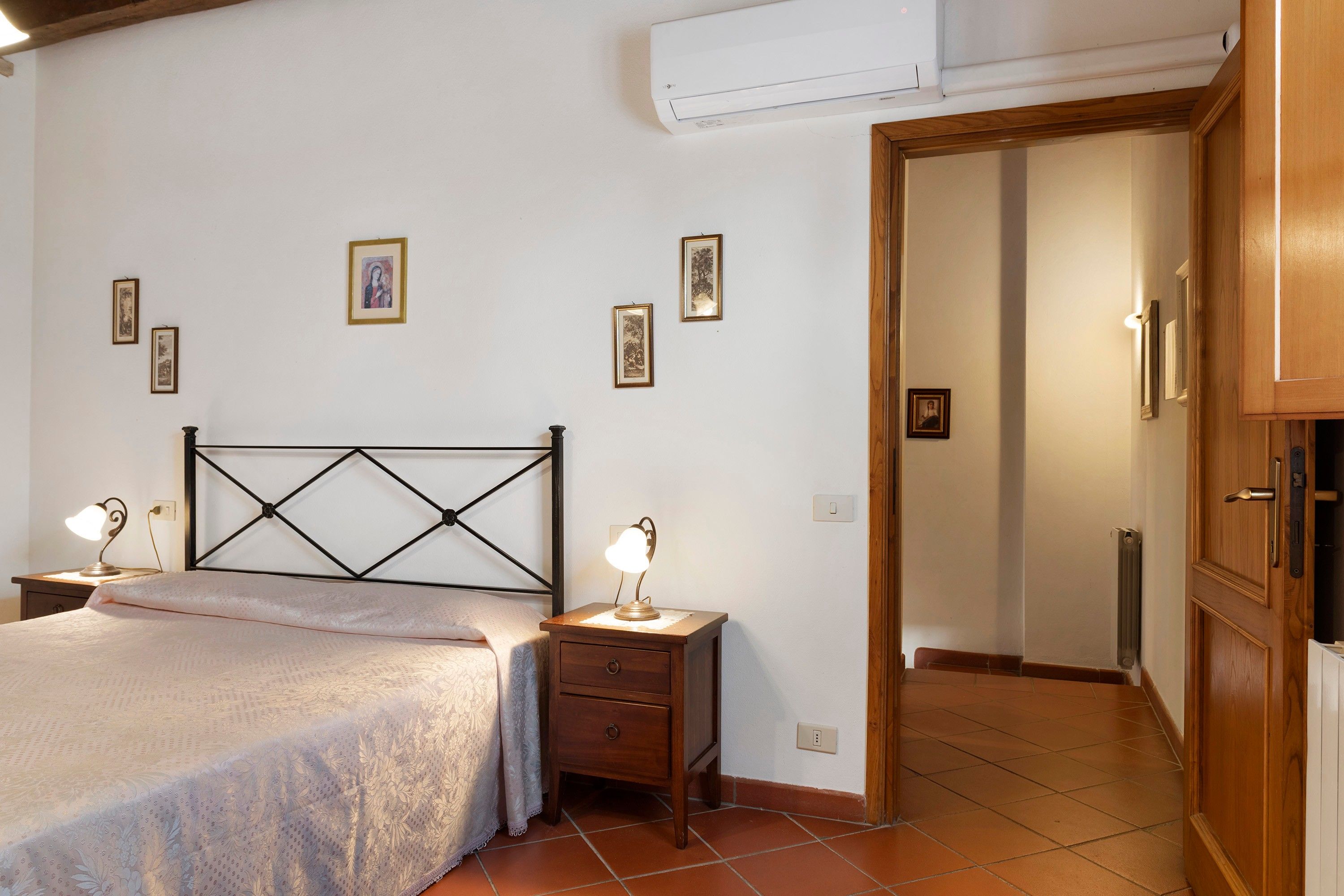 Quarto com cama, mesas de cabeceira, guarda-roupa de madeira e uma porta que dá para outro cômodo.