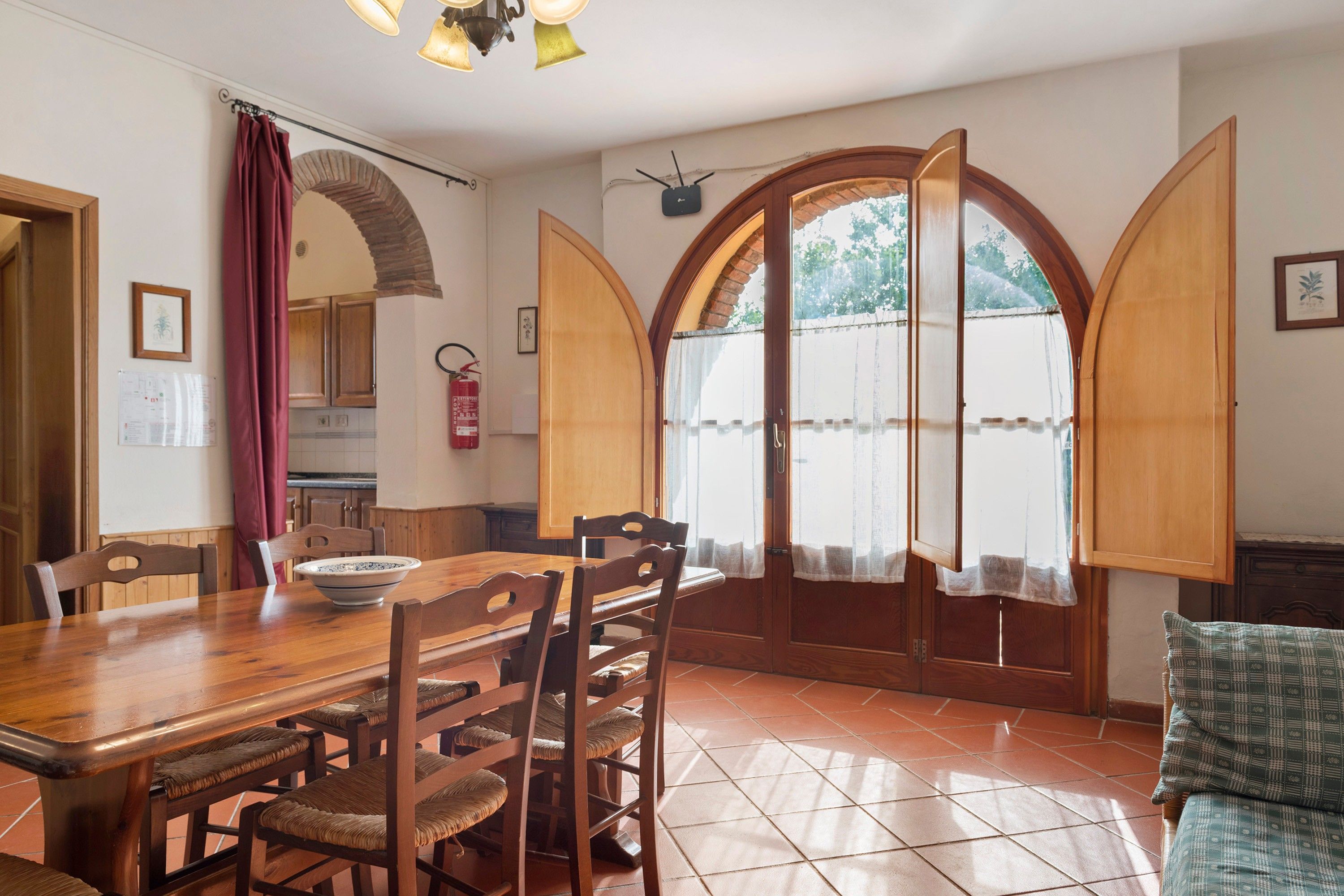 A dining area with a wooden table and chairs, a fire extinguisher, and a view through an arched window.