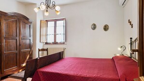A bedroom with a wooden wardrobe, a bed with a red bedspread, a window with curtains, and a wall-mounted air conditioning unit.