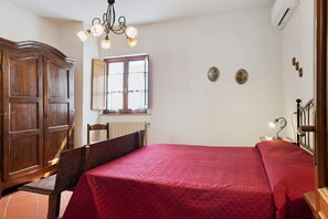 A bedroom with a wooden wardrobe, a bed with a red bedspread, a window with curtains, and a wall-mounted air conditioning unit.
