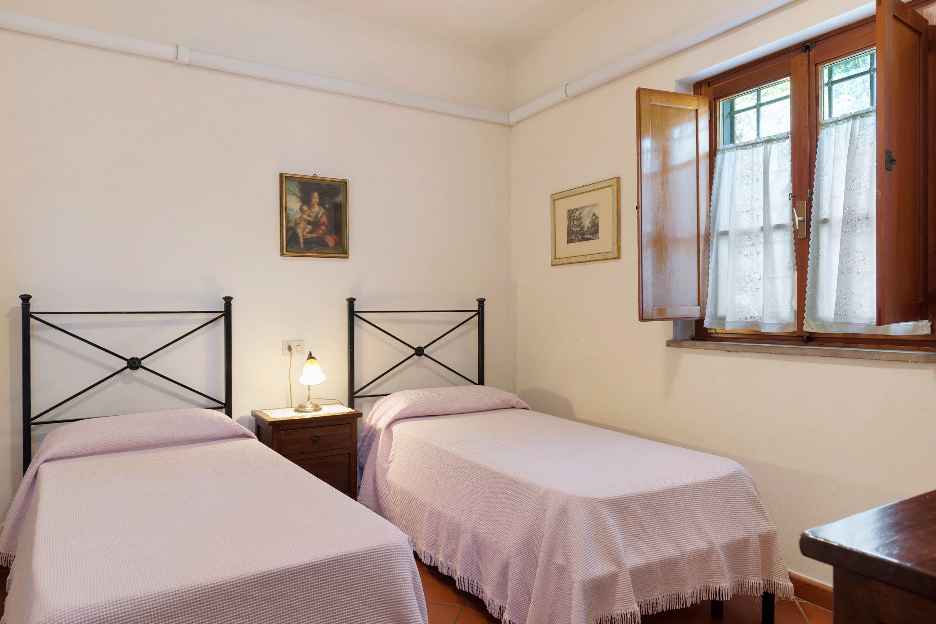 A bedroom with two single beds, a nightstand with a lamp, a window with curtains, and two framed pictures on the wall.