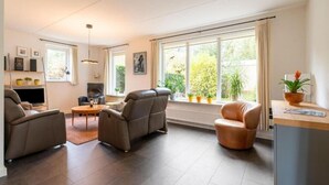 A modern living room with a sofa, armchairs, a coffee table, and a TV. There are potted plants and a window with a view of greenery. - Comfortable country house in Drenthe (Schoonloo)