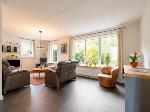 Ein modernes Wohnzimmer mit einer Couch, Sesseln, einem Couchtisch und einem Fernseher. Üppige Zimmerpflanzen und ein Fenster mit Blick auf Grün.