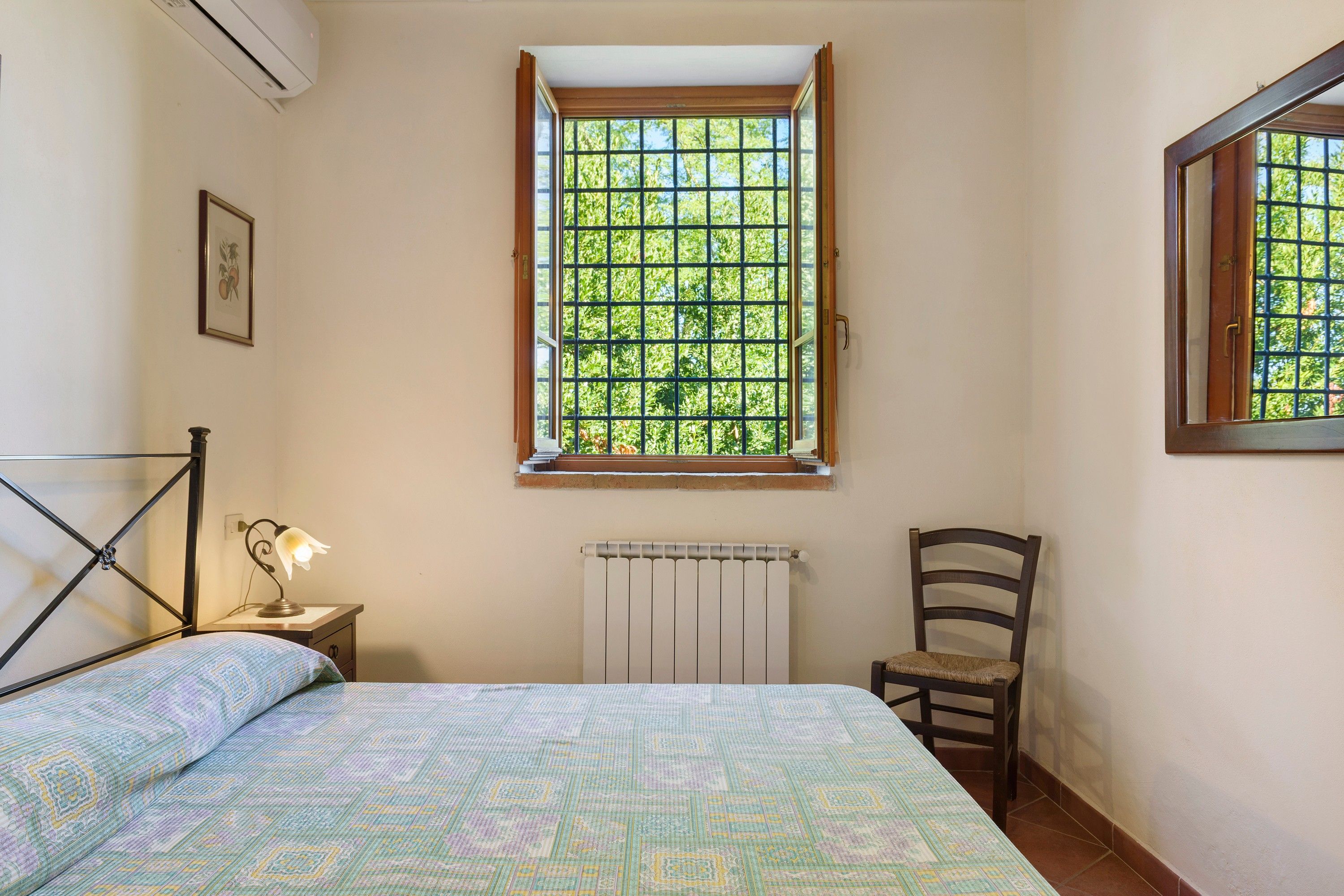 Un dormitorio con cama, una silla, un radiador y una ventana con vistas a la vegetación.