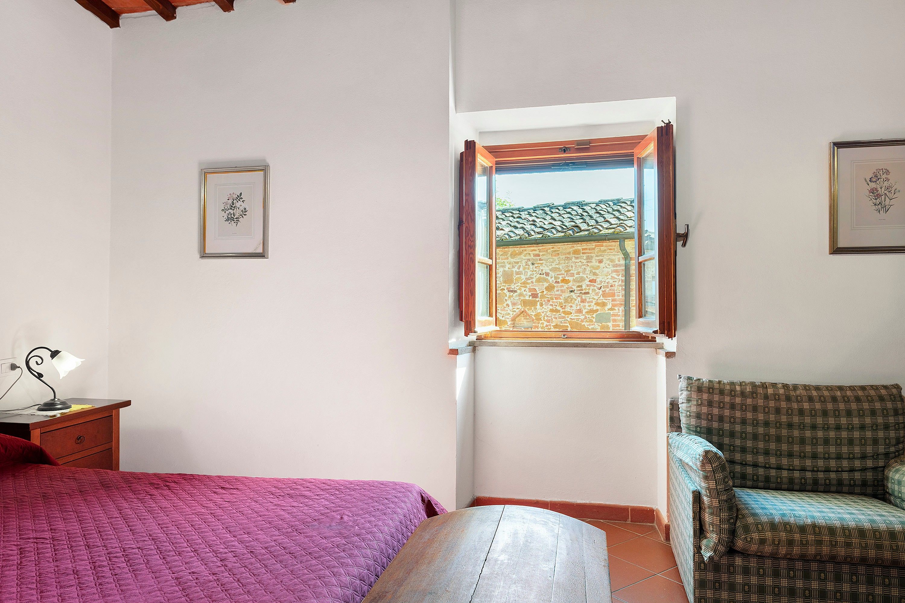 Ein Schlafzimmer mit einem Bett, einem Schreibtisch mit Lampe, einem Fenster mit Blick auf eine Ziegelwand und einem gemusterten Sessel.