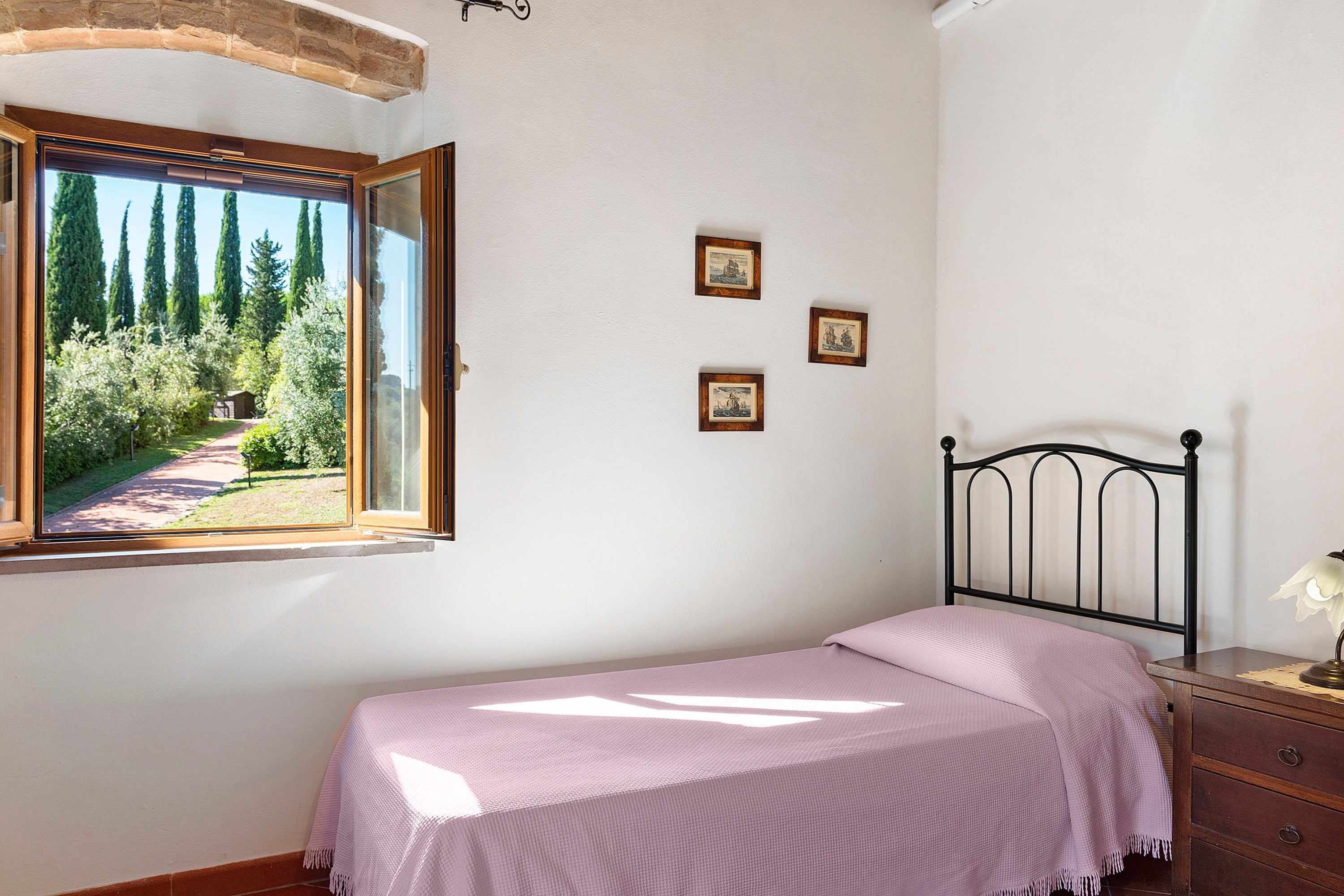 Un dormitorio con cama, mesita de noche, lámpara y ventana con vista a árboles y un sendero.