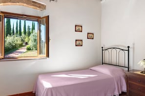 A bedroom with a bed, a nightstand, a lamp, and a window with a view of trees and a path. - Michelangelo Apartment: Panoramic Views & Spacious Comfort (Montaione)